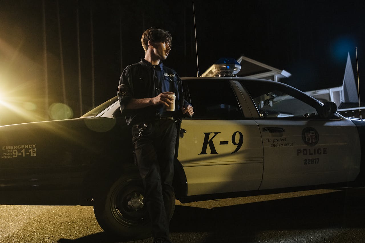 A police officer stands by a K-9 unit car at night, holding a coffee, illuminated by streetlights.