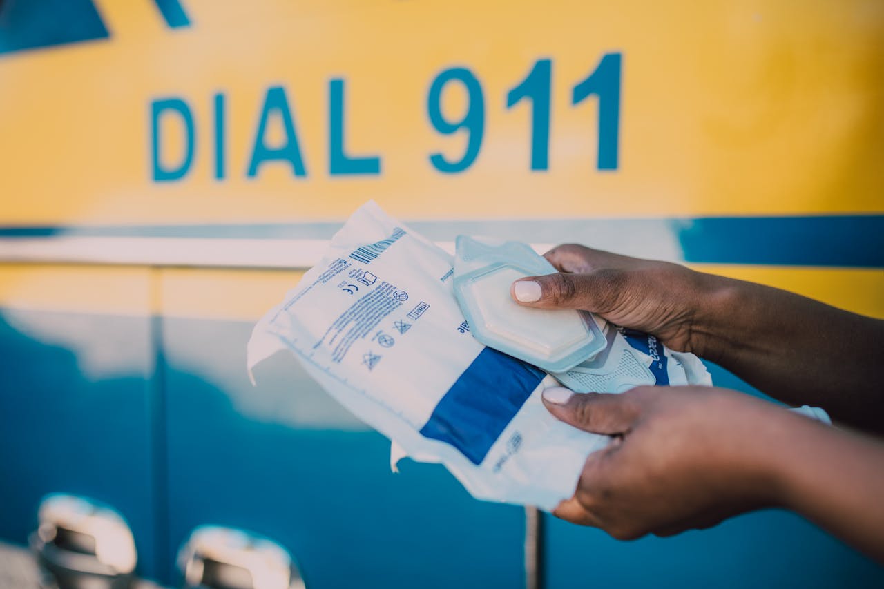 Hands holding medical supplies in front of an ambulance with 'Dial 911' sign.